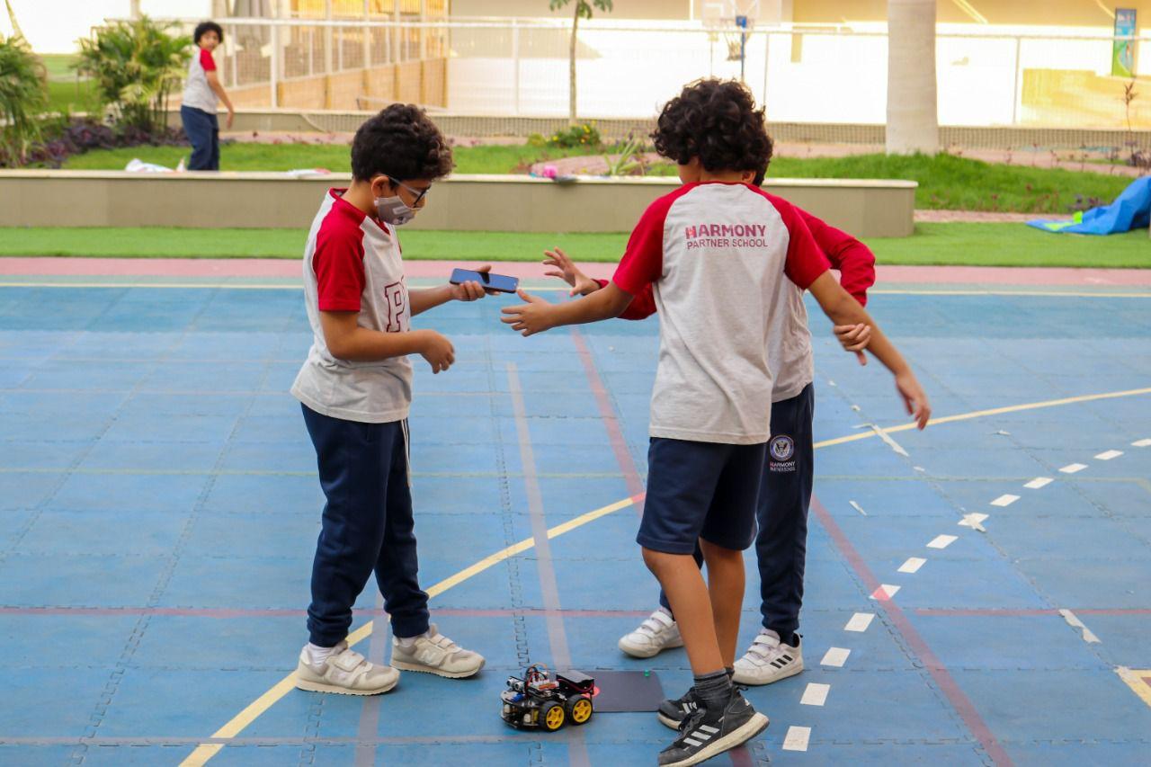 Two children from IVY STEM International School wearing uniforms, using a robotic vehicle in the schoolyard. They exchange a tablet or remote control device, engaging in educational activities during the COVID-19 pandemic.
